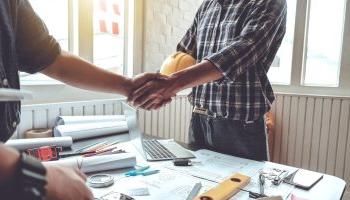 Two People Shaking Hands Over A Desk With A Computer, Blueprints, And Other Planning Items.